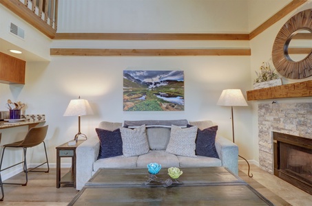 A living room with a beige sofa, four cushions, two lamps, a framed landscape photo above the sofa, a fireplace, a round mirror, a table with decorations, and a small bar area with two stools.