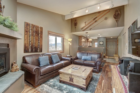 A cozy living room with leather couches, rustic decor, wooden ceiling beams, and a mix of modern lighting. There's a fireplace, a wall-mounted painting, and snowshoes and skis mounted on the walls.