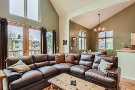 A spacious living room with high ceilings, a sectional sofa, large windows, a dining area, and a chandelier. A coffee table with a remote control is in the center.