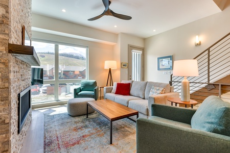 Contemporary living room with a large window, two green armchairs, a grey sofa with a red pillow, two lamps, a coffee table, a ceiling fan, and a wall-mounted TV above a stone fireplace.