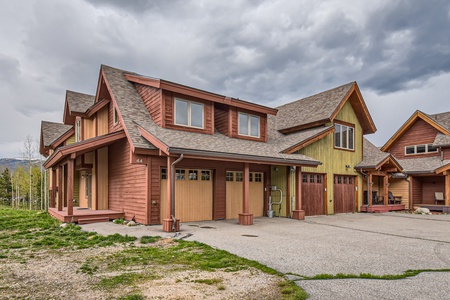 A two-story house with wooden exterior, three garage doors, and a porch, set against a backdrop of cloudy skies. The house has a mix of brown and green tones.