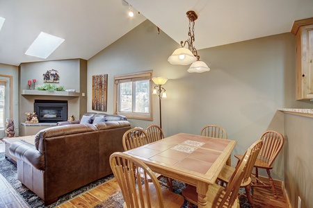 A cozy living and dining area with a brown leather couch, a wooden dining table with chairs, a fireplace, and skylights. The room features tan walls and natural light from a window.