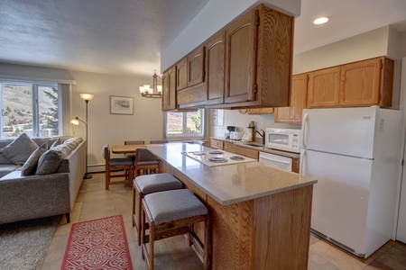 A kitchen with wooden cabinets, an electric stove, a microwave, and a refrigerator. There is a countertop with stools, a dining table, and a seating area with a sofa in the background.