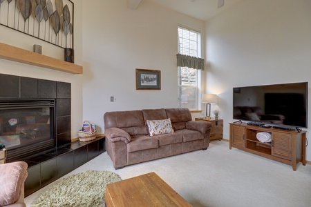 Living room with a brown sofa, wooden coffee table, flat-screen TV on a wooden stand, fireplace, and window with blinds and valance. Walls display framed art. Light colors dominate the decor.