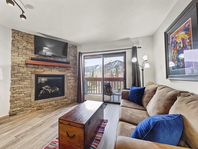 A cozy living room with a stone fireplace, wall-mounted TV, brown sofa, and wooden chest. A sliding glass door opens to a balcony with mountain views. A vibrant painting and floor lamp decorate the space.