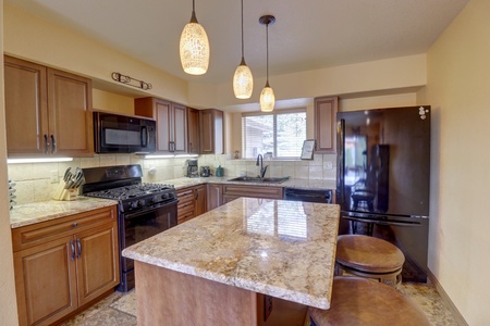 A modern kitchen featuring wooden cabinetry, granite countertops, a center island with two stools, pendant lights, and black appliances including a refrigerator and a gas stove. A window provides natural light.