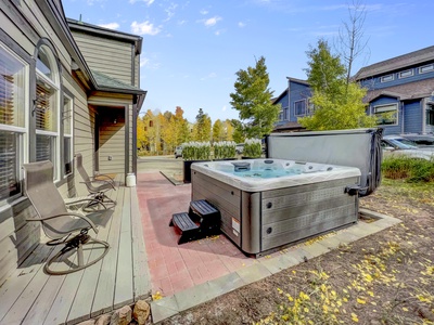 A backyard with a hot tub on a brick patio, two chairs, and a wooden house exterior, surrounded by trees and additional houses in the background under a blue sky.