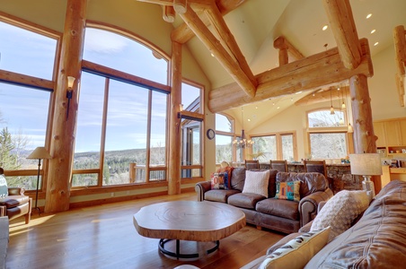 Spacious living room with large windows, log beams, leather couches, and a rustic coffee table. The room offers a view of a forested landscape outside and a modern kitchen in the background.