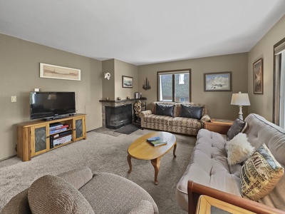 A living room with a couch, loveseat, and an upholstered chair. A wooden coffee table is in the center. A flat-screen TV sits on a stand. A fireplace is in one corner. Artwork hangs on the walls.