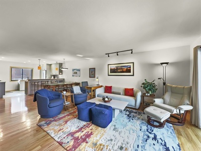 A modern living room with a light-colored sofa, blue and red accent chairs, a patterned rug, a floor lamp, and a wall-mounted framed artwork. The open kitchen and dining area are visible in the background.