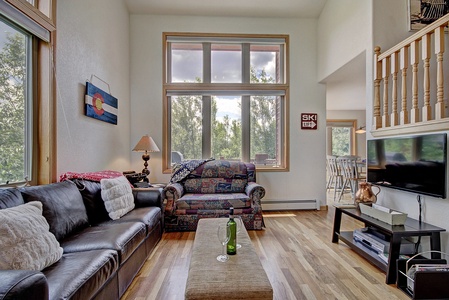 A living room with large windows, a black leather couch, a patterned armchair, a small table with a drink, and a television. An adjacent room with a dining area is partially visible.