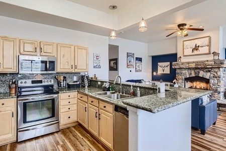 A modern kitchen with light wood cabinets, granite countertops, stainless steel appliances, and a double sink, adjacent to a living area with a stone fireplace. Warm, cozy and inviting space.