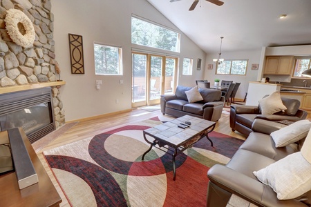 A bright living room featuring a stone fireplace, leather sofas, a patterned rug, and a coffee table. Large windows and a ceiling fan complement the space with an open view of the outdoors.