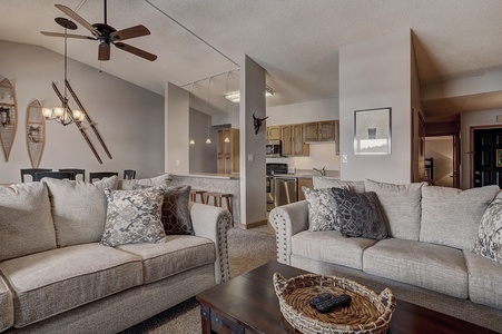 An open-plan living room with beige sofas, a wooden coffee table, and a kitchen in the background featuring wooden cabinets and appliances. Ceiling fans and unique wall decor add to the space.