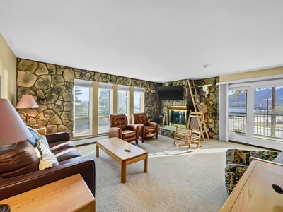 A living room with stone walls, a large window, a brown leather couch, two brown armchairs, a wooden rocking chair, a coffee table, and a TV mounted above a fireplace.