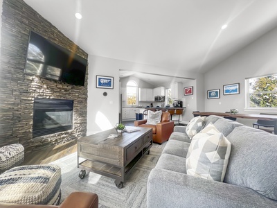 A living room with a stone fireplace, TV above it, gray couch, chair, ottoman, wooden coffee table, and view into a white kitchen.