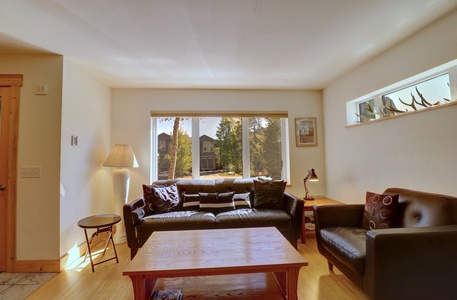 A living room with a large window, sunlight pouring in, black sofas, wooden coffee table, floor lamp, side tables, and wall shelves with decorative items.