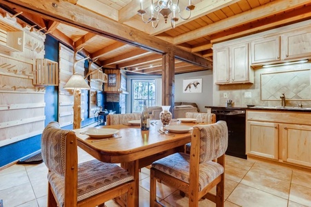 A cozy dining area with a wooden table and four chairs is set against a background of exposed wooden beams and a small kitchen. There's a living area with a fireplace and large windows nearby.