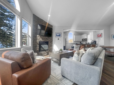 A modern living room with a stone fireplace, wall-mounted TV, large windows, gray sofa, and brown leather armchair. An open kitchen with white cabinets and a wooden dining table is visible in the background.