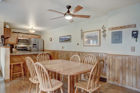 A kitchen and dining area with wooden furniture, including a dining table with six chairs. The kitchen has a refrigerator, stove, and cabinets. Ceiling fan and framed pictures on the wall are visible.