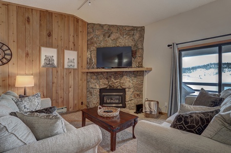 A cozy living room with a stone fireplace, mounted TV, and light gray sofas. The room has wooden walls, a coffee table, and large windows showcasing a snowy outdoor view.