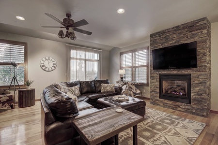 A cozy living room with a brown leather sectional sofa, stone fireplace with a mounted TV above, rustic wooden furniture, a clock on the wall, large windows with blinds, and a ceiling fan.