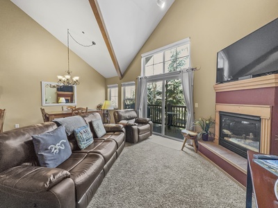 A cozy living room with vaulted ceilings, leather sofas, a fireplace, large windows with curtains, and a TV. Patio doors lead to a balcony with greenery outside. A chandelier hangs above a dining area.