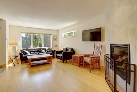 A spacious living room with wooden floor, leather sofas, a coffee table, armchairs, a wall-mounted TV, a fireplace with a screen, a standing lamp, and large windows letting in natural light.