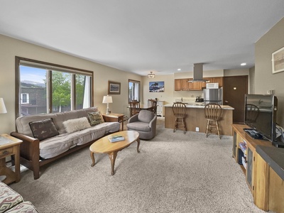 A cozy living room with a futon, armchair, wooden coffee table, and TV. Open kitchen with wooden bar stools, light carpet, and large windows allowing natural light. Wall art and lamps are present.