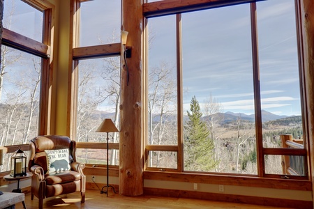 A spacious room with large windows showcasing a scenic mountain and forest view. The room has a leather armchair with a pillow, a floor lamp, and wooden beams framing the windows.