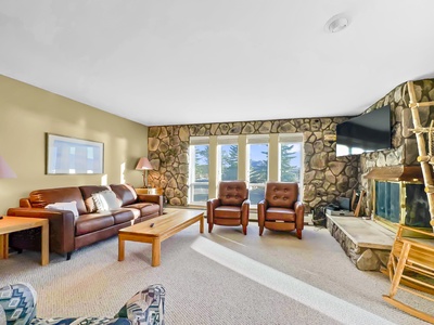 A cozy living room with a brown leather sofa, two armchairs, a wooden coffee table, and a stone fireplace on the right. Large windows on the back wall let in natural light. A TV is mounted above the fireplace.