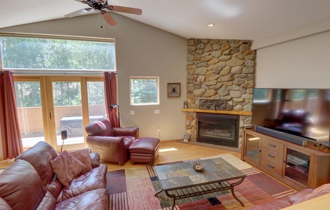 A living room with a stone fireplace, large TV, leather sofas, and armchair. Sliding glass doors and windows provide natural light. The room has a wooden floor and colorful area rug.