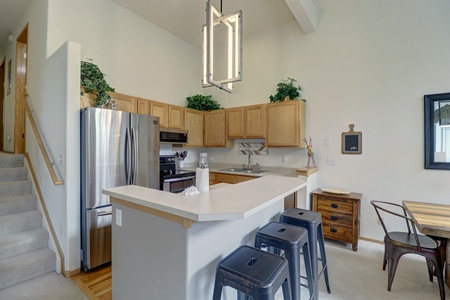 A modern kitchen with wooden cabinets, stainless steel appliances, a counter with three dark stools, and a small dining area with table and chairs. A staircase is visible to the left.