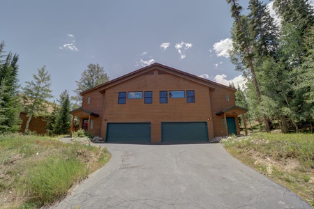 A two-story brown wooden house with two green garage doors, a paved driveway, and surrounded by trees on a sunny day.