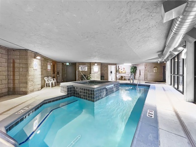 Indoor swimming pool area with a hot tub, surrounded by a tiled floor. A few plastic chairs are placed along one wall, and signs with rules are posted. Large windows provide natural light.