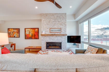 A cozy living room with a stone fireplace, abstract paintings on one wall, a flat-screen TV, and large windows offering an outside view. A ceiling fan is visible above.