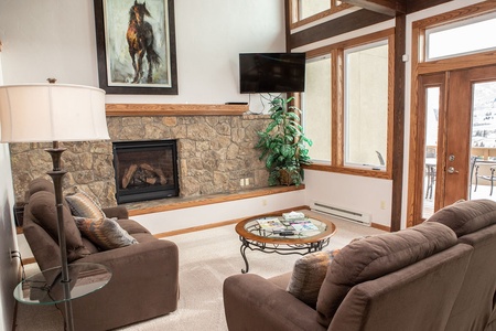 A cozy living room with a stone fireplace, two brown sofas, a round glass coffee table, a flat-screen TV, a large painting of a horse, a floor lamp, and a plant. Large windows offer outdoor views.