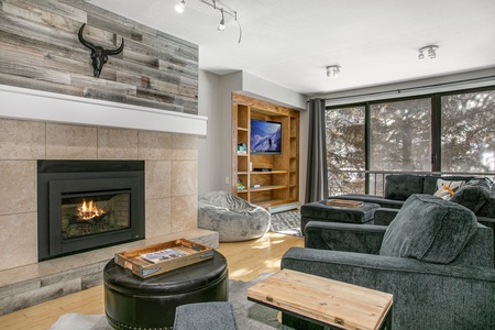 A cozy living room with a lit fireplace, gray armchairs, a wooden shelf with a television, a large window, and natural light. The wall above the fireplace has a bull skull decor.