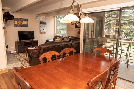 A cozy dining room with a wooden table and chairs, adjacent to a living area with a leather sofa, a TV, and sliding glass doors leading to an outdoor balcony.