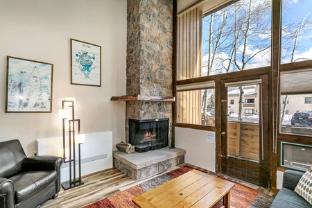 A cozy living room with a stone fireplace, leather armchair, modern floor lamp, wooden coffee table, and large window offering a view of snowy trees and houses outside.