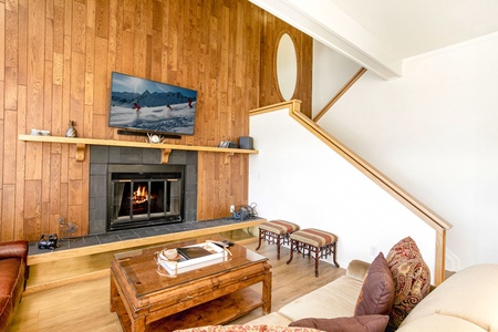 Living room with a wooden accent wall, fireplace, and a TV mounted above. The area includes a coffee table, couch, and two stools. An open staircase runs adjacent to the setup.