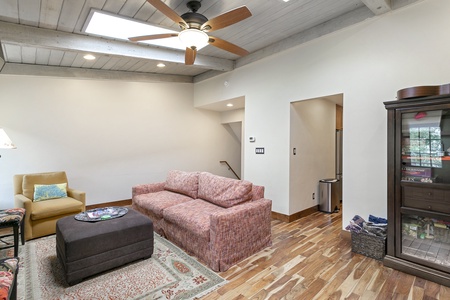 Living room with a ceiling fan, two couches, a yellow chair, an ottoman, and a cabinet. There is a rug on wood flooring and a staircase in the background.