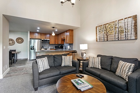 A modern living room featuring two gray sofas with patterned cushions, a wooden coffee table, and a kitchen area in the background with wooden cabinets and stainless steel appliances.