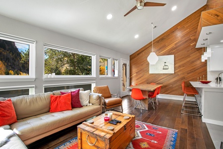 A modern, open-concept living and dining area featuring a white couch with red pillows, a wooden coffee table, and an adjacent dining space with orange chairs and a wooden accent wall.