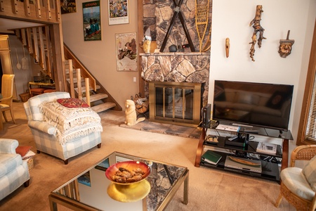 A cozy living room with a stone fireplace, TV, armchairs, and a glass coffee table. Stairs lead to an upper level, and the walls are adorned with various posters and masks.