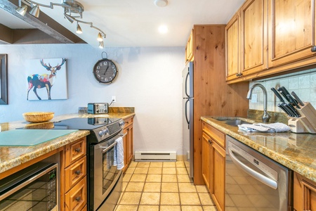 A kitchen with wooden cabinets, stainless steel appliances, and granite countertops. A clock and colorful moose painting adorn the walls. Utensils and a cutting board are placed on the counter.