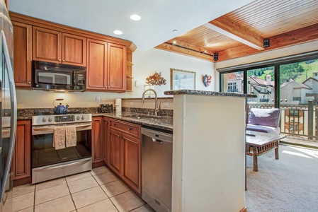 Modern kitchen with wooden cabinets, stainless steel appliances, and granite countertops. The kitchen opens to a living area with a large window and balcony, featuring mountain views.