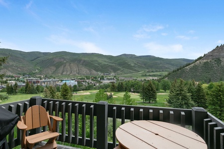 View from a balcony overlooking a green valley with mountains in the background. The balcony has wooden chairs and a round table. A small town is visible in the distance.