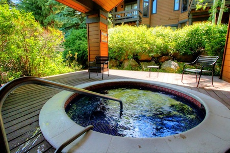 Outdoor hot tub surrounded by lush greenery and a wooden deck, with two black chairs positioned nearby.