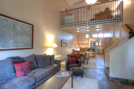 A modern living room with a gray couch, armchair, coffee table, and wall artwork. A loft area is seen above, and a dining table and kitchen are in the background. Stairs lead to the loft.